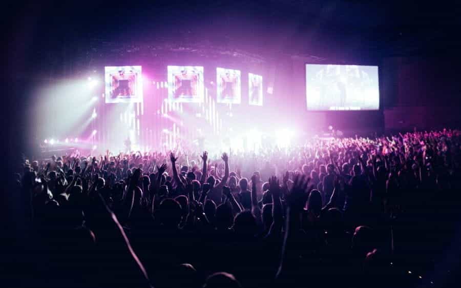 dramatic and bright concert stage seen from the back of a stadium, where silhouettes of fans scream with their arms up.