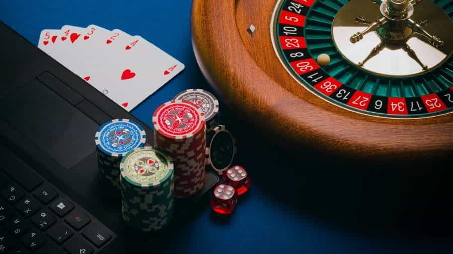 A roulette wheel, a stack of cards, and poker chips sit next to an open black laptop.