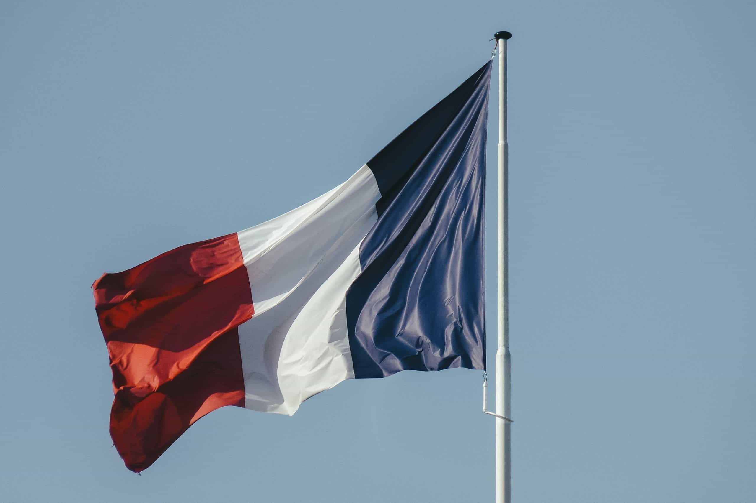 Blue, white, and red French flag on a flagpole with blue sky.