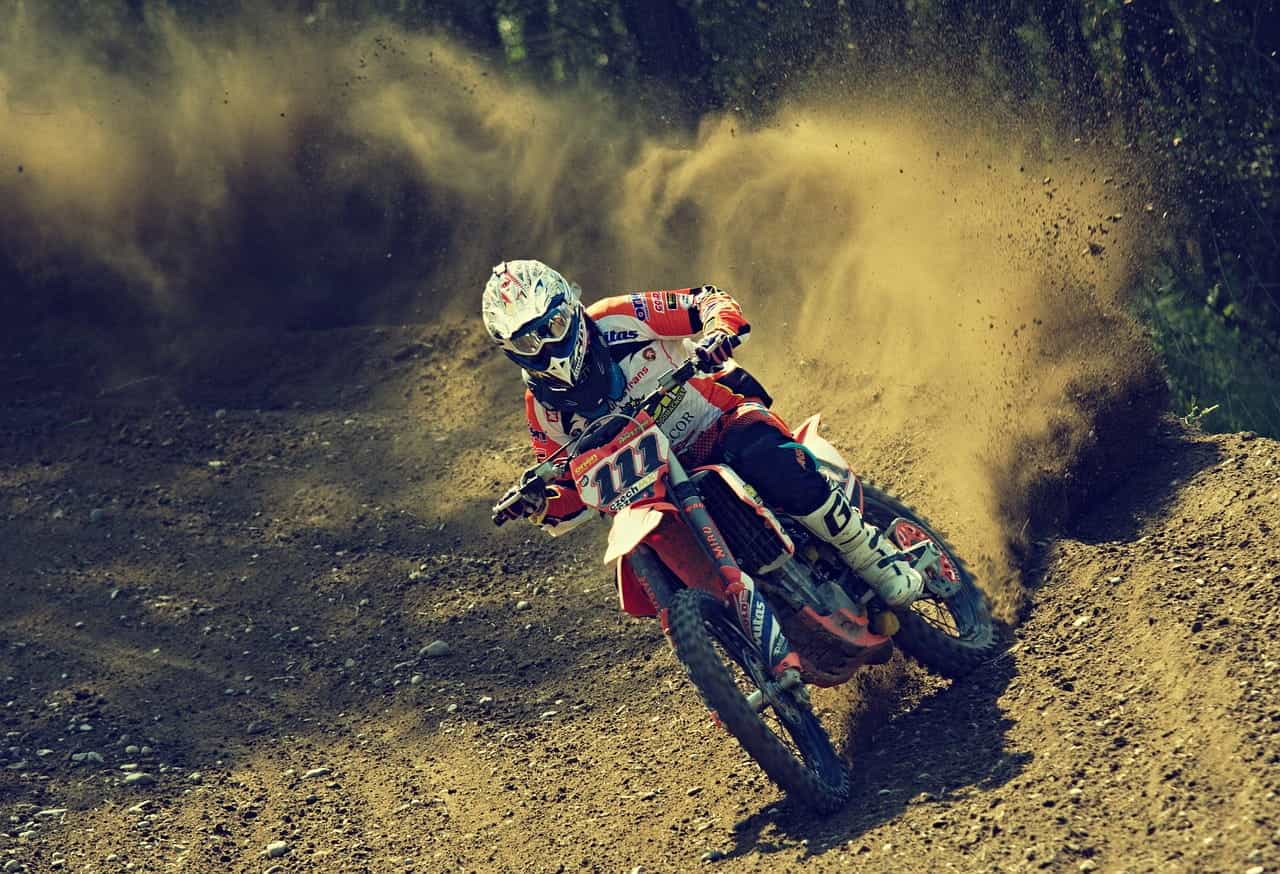 A person in full racing gear riding a dirt bike/motorcycle during a race, with a fountain of dirt being kicked up behind the back wheel of the motorcycle.