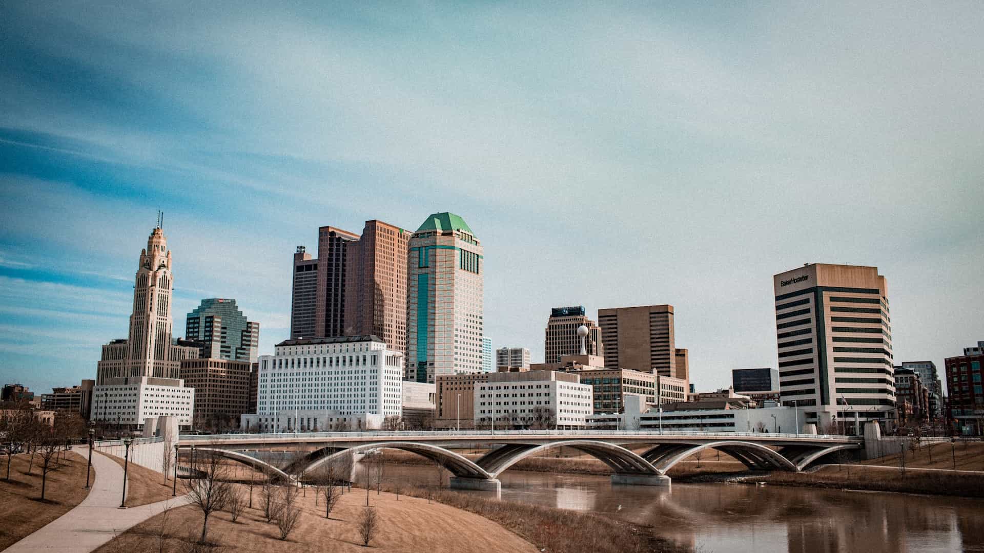 The dense downtown district of Columbus, Ohio, featuring several tall buildings and skyscrapers and a small bridge cutting across a river.