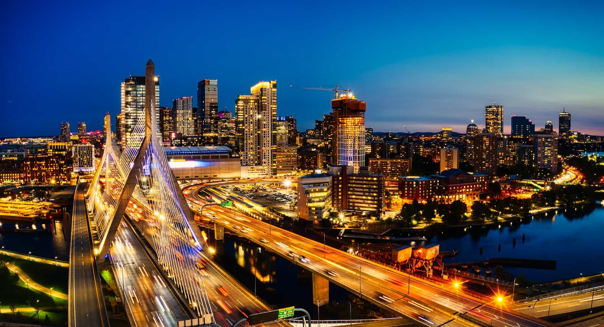 The towering skyline of the city of Boston, Massachusetts at dusk, featuring several skyscrapers and bridges cutting across the Charles River.